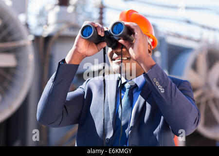 African electrician engineer using binoculars looking at electricity substation Stock Photo