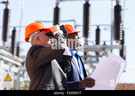 middle aged engineer and colleague with binoculars on electrical substation Stock Photo