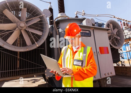 experienced senior electrical technician using laptop computer in front of transformer Stock Photo