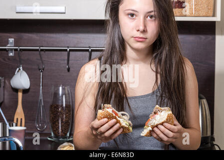 Teenaged girl is eating burger in kitchen interior. Stock Photo