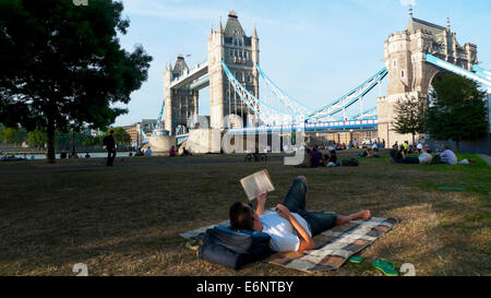 Man lying on blanket reading a book on a summer evening in park by River Thames with a view of Tower Bridge Central London England UK  KATHY DEWITT Stock Photo