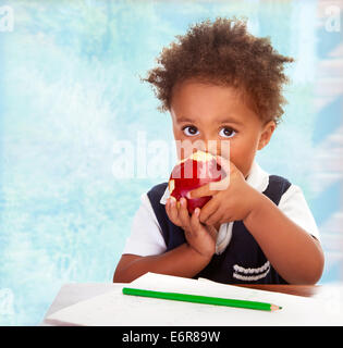 Portrait of cute little African boy sit behind a desk and biting big red apple, drawing using green pencil, having lunch i Stock Photo