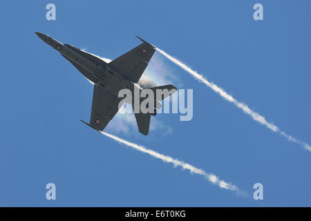 A Swiss Air Force McDonnell Douglas F/A-18C Hornet in silhouette creates a rainbow and vapour trail against a blue sky. Stock Photo