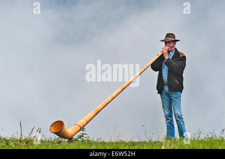 An alphorn player, playing his instrument on a ridge above the alpine toen of Nendaz. Cloudy background of mountains. Stock Photo