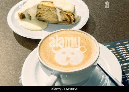 Cappuccino with little bear decoration drawn into the foam. In the background the austrian specialty "Apfelstrudel" with vanilla Stock Photo