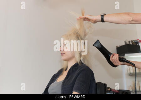 Beautiful blonde girl sitting, getting hair dried. Hands of hairdresser visible, holding blow dryer Stock Photo