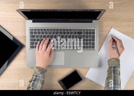 Female programmer writing notes on piece of paper while typing laptop keyboard, top view. Stock Photo