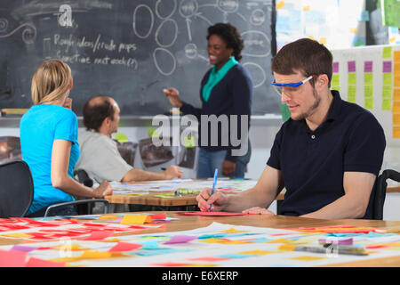 Engineering instructor working on flowchart on blackboard while students working on project one man with spinal cord injury and other one with Aspergers Stock Photo