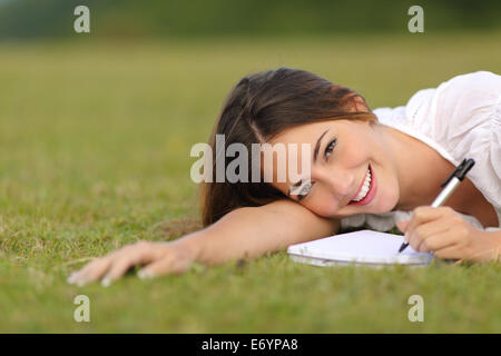 Happy woman lying on the grass and writing in a notebook with unfocused green background Stock Photo