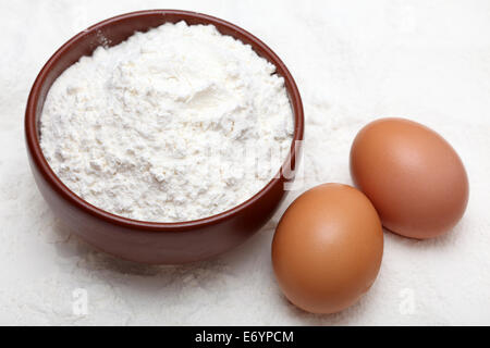 Flour in bowl and two eggs on flour background. Closeup. Stock Photo