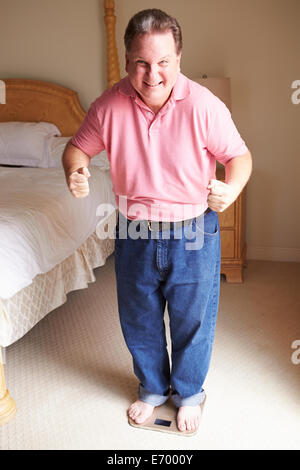 Happy Overweight Man Standing On Scales In Bedroom Stock Photo