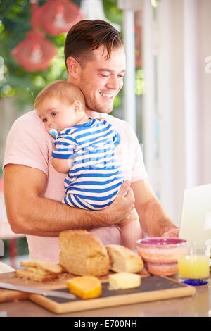 Father Holding Baby And Making Snack Whilst Using Laptop Stock Photo