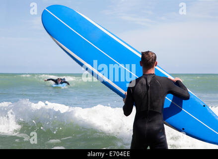 Rear view of male surfer with surfboard watching friend surfing Stock Photo