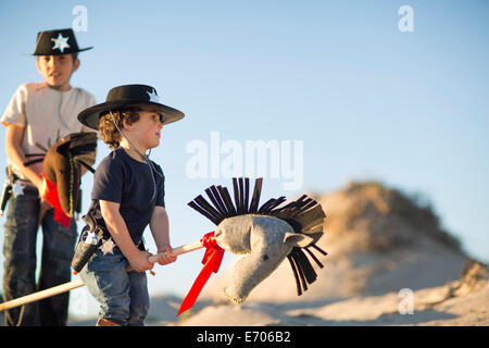 Two brothers dressed as cowboys with hobby horses in sand dunes Stock Photo
