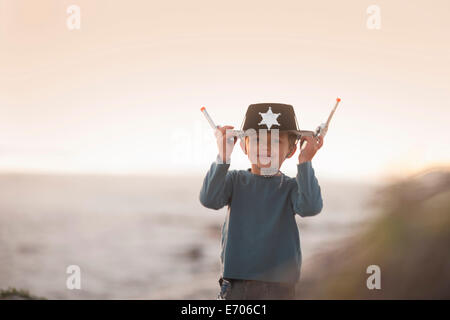 Boy dressed as cowboy sheriff holding onto hat and toy guns in sand dunes Stock Photo