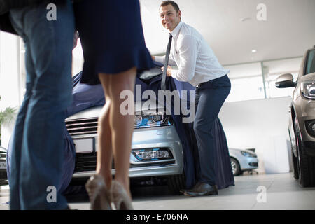 Low angle view of salesman uncovering new car for mid adult couple in car dealership Stock Photo