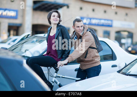 Young woman sitting on boyfriend's bicycle handlebar moving through city traffic jam Stock Photo