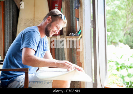 Young male surfer sitting in shed waxing surfboard Stock Photo