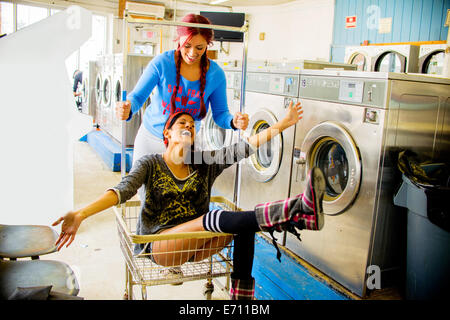Two young women in laundromat, one pushing the other along in trolley Stock Photo