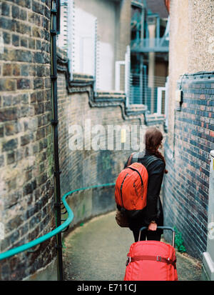 A woman walling down a narrow street, pulling a suitcase and holdin an orange backpack. Stock Photo