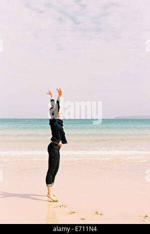 A woman standing barefoot on the sand raising her arms above her head, in a gesture. Stock Photo