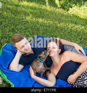 A couple sitting on a picnic rug. A small dog licking the face of a woman. Stock Photo