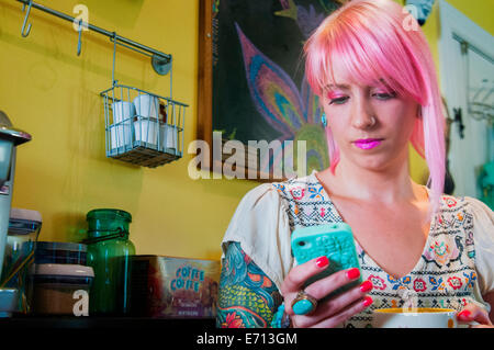 Young woman with pink hair reading text message on smartphone in kitchen Stock Photo
