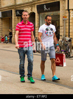 Two young men of different racial origins wearing casual clothes, one with beard and carrying shopping bag, walking along street Stock Photo