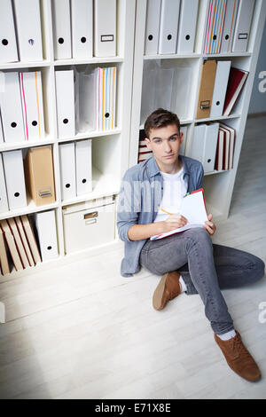 Serious student sitting on the floor and making notes in copybook Stock Photo