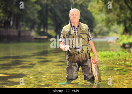 Mature fisherman holding a fish and fishing rod in a river Stock Photo