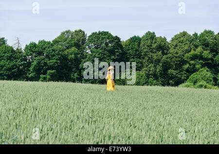 Beautiful farmer woman in yellow dress and hat walk in agriculture wheat field and strong wind blow. Stock Photo