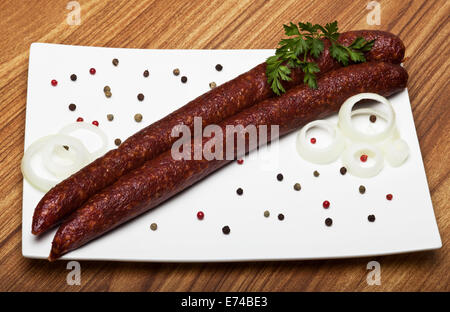 Two sausages surrounded with black green and red pepperbeans slices of onion and parsley on a white plate. Wooden background. Stock Photo