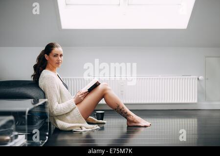 Beautiful brunette woman reading a book by a sofa. Attractive female model sitting on floor with a book looking at camera. Stock Photo