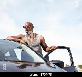 Portrait of smart young man leaning on car. African male model wearing sunglasses standing by his car looking away with door. Stock Photo