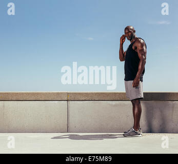 Portrait of fit young man standing on a walkway looking at camera. African fitness model wearing earphones. Stock Photo