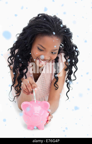 Composite image of a young girl lying on the floor putting money into a piggy bank Stock Photo