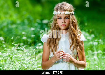 Portrait of cute blond girl dressed in white standing in green field. Stock Photo
