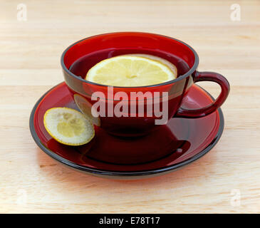 A cup of lemon tea with slices floating in the water and a small piece of rind on the saucer atop a wood counter top. Stock Photo