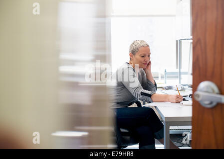 A woman working in an office alone. Focusing on a task, making notes with a pencil. Stock Photo