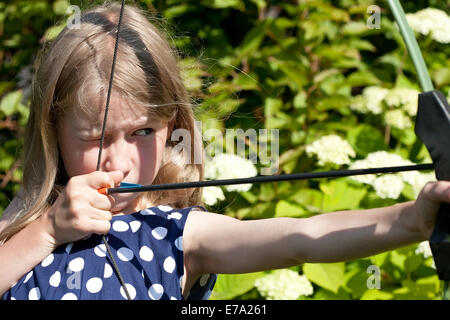 little caucasian girl aiming arrow from big bow on outdoor nature background Stock Photo