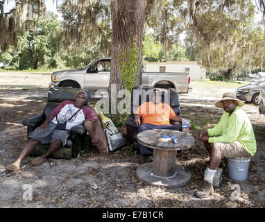 three black men sitting under a tree.  Two are sitting in recliners and one on a bucket. Stock Photo