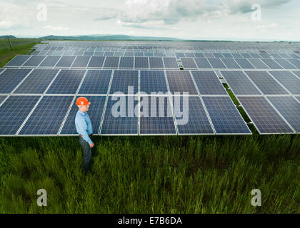 man dressed in engineer clothes and equipment standing near solar panels, outdoors, with clouded sky Stock Photo