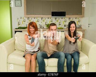 three young happy caucasian people sitting on couch, having fun while playing video game Stock Photo