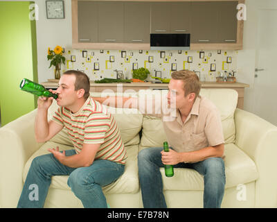 two young caucasian men sitting on couch, holding beer bottles, consolation Stock Photo