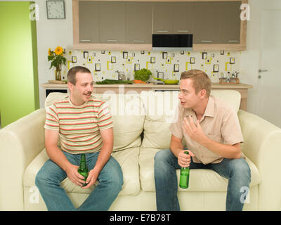 two young caucasian men sitting on couch, holding bottles of beers and having conversation Stock Photo