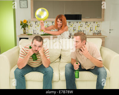 two caucasian men sitting on couch holding beer bottles, having conversation, woman in background trying to hit one of them with Stock Photo
