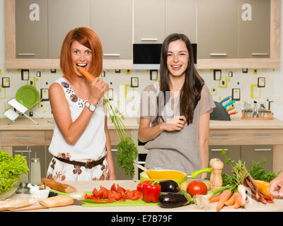 portrait of two young beautiful caucasian women standing in kitchen and laughing, one of them biting carrot Stock Photo