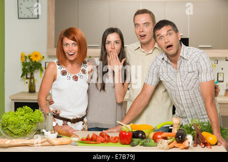 portrait of four young caucasian people standing in kitchen, table with colorful mixed vegetables, acting surprised Stock Photo