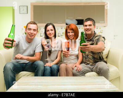 friends sitting at home on sofa, making a toast with drinks and watching tv Stock Photo