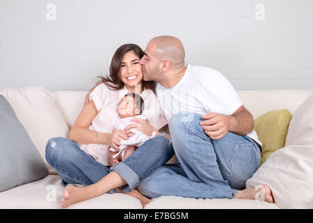 Handsome young father kissing beautiful smiling mother with cute newborn baby on hands, having fun together at home Stock Photo